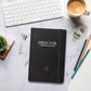 Charcoal matte hardcover notebook on a desk beside a keyboard, coffee cup, and pens.